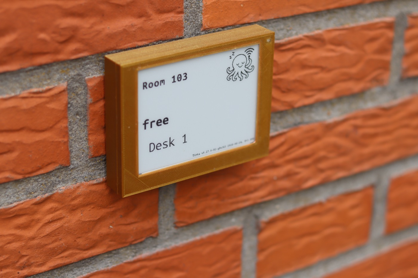Tinta display mounted on a brick wall, showing a room, free status, and desk number